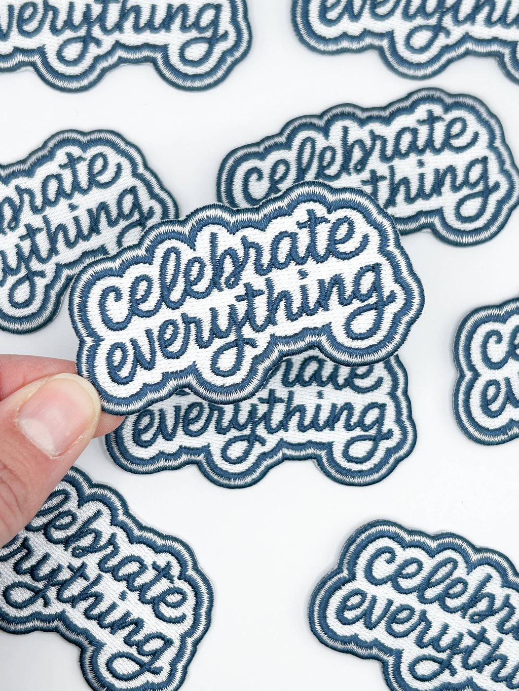 Blue embroidered patch with 'Celebrate Everything' text held over a sheet of similar patches.