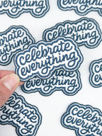 Blue embroidered patch with 'Celebrate Everything' text held over a sheet of similar patches.