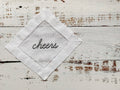 A white embroidered cotton handkerchief folded into a diamond shape, featuring the word 'cheers' stitched in black script in the center, displayed on a rustic white-washed wooden surface.