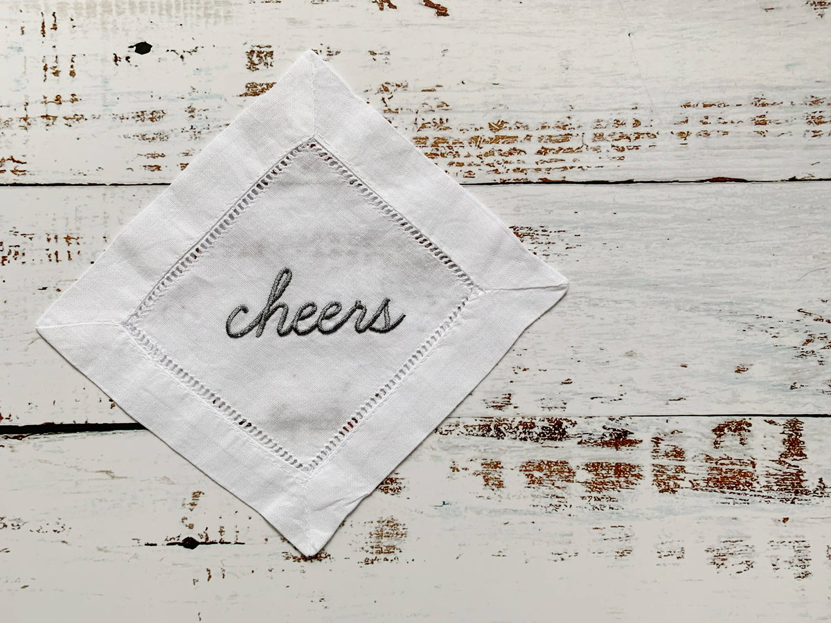 A white embroidered cotton handkerchief folded into a diamond shape, featuring the word 'cheers' stitched in black script in the center, displayed on a rustic white-washed wooden surface.