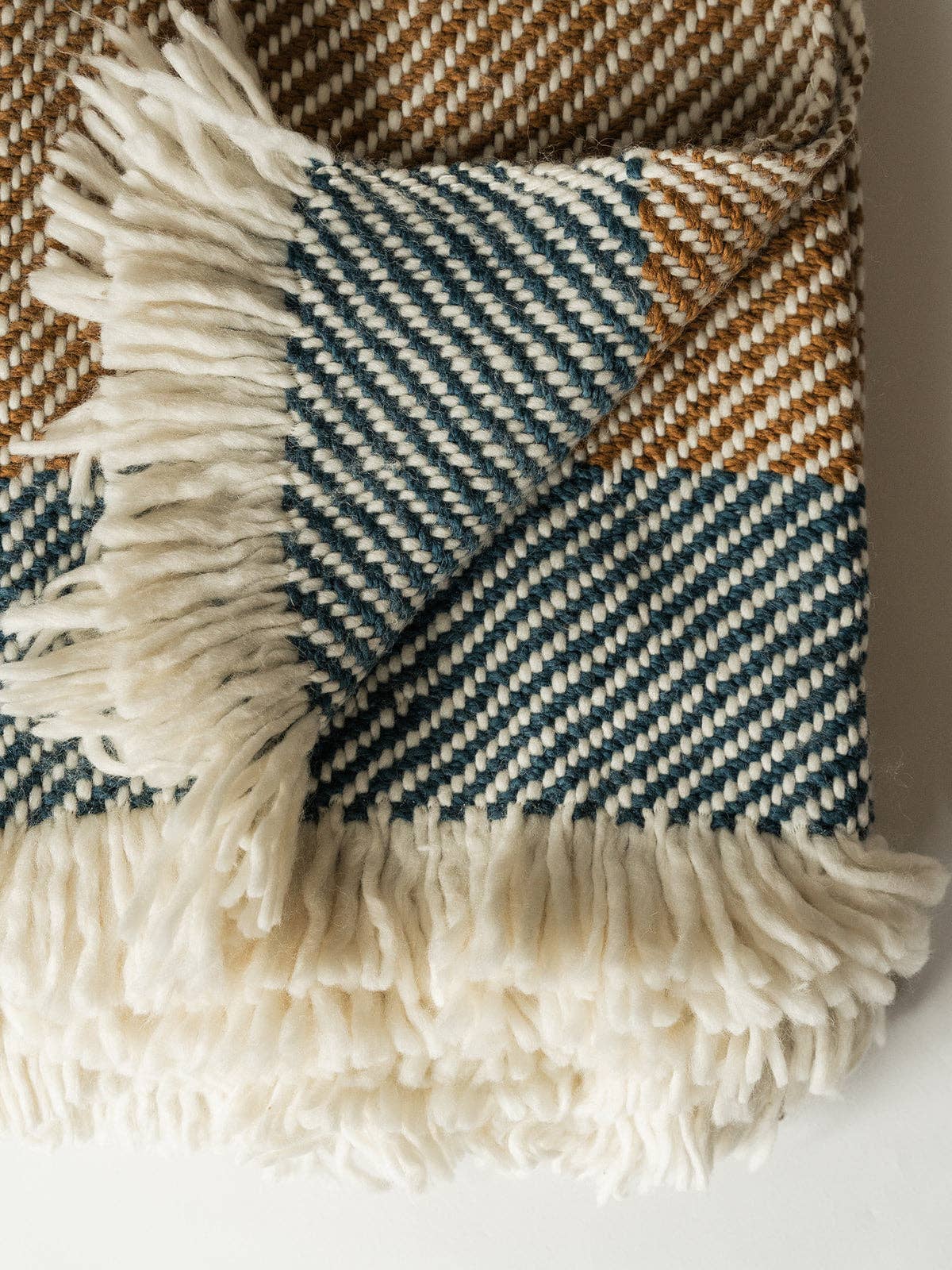A close-up of a woven throw blanket with fringe, featuring a color-blocked pattern of cream, slate blue, and tan herringbone and diagonal stripe textures.