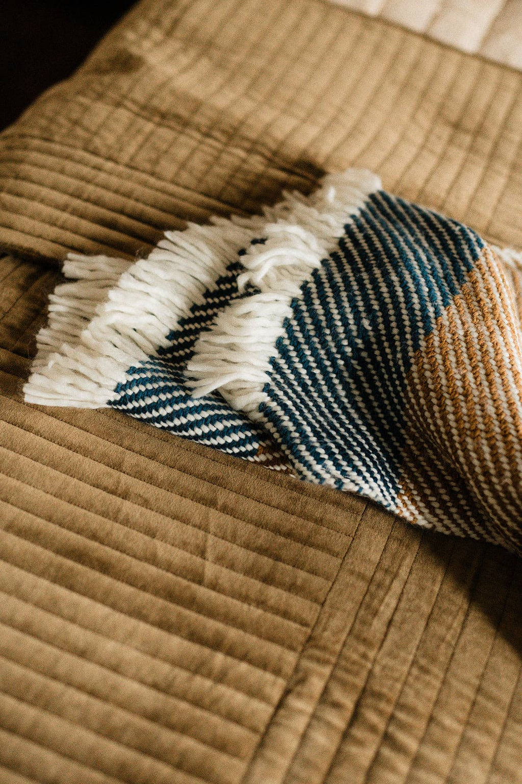 A close-up of a folded woven throw blanket with white fringe, featuring stripes of navy blue, teal, cream, and tan, resting on a brown quilted blanket.
