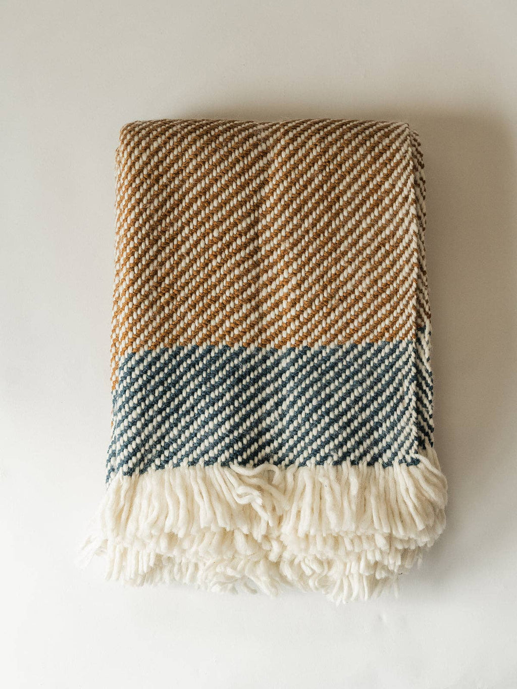 A folded woven throw blanket with a herringbone pattern, featuring a tan and cream upper section, a muted blue-gray middle band, and a thick cream fringe at the bottom, displayed against a plain off-white background.