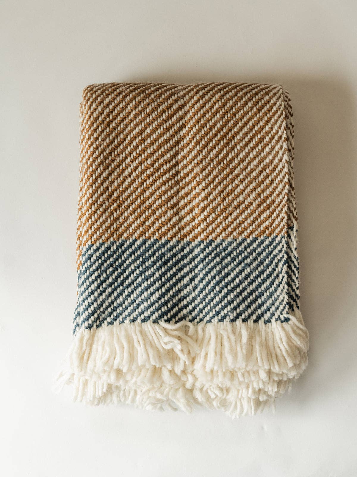 A folded woven throw blanket with a herringbone pattern, featuring a tan and cream upper section, a muted blue-gray middle band, and a thick cream fringe at the bottom, displayed against a plain off-white background.