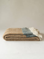 A folded woven throw blanket with a herringbone pattern in beige, brown, and muted blue-gray, finished with white fringe tassels, set against a plain light beige background.