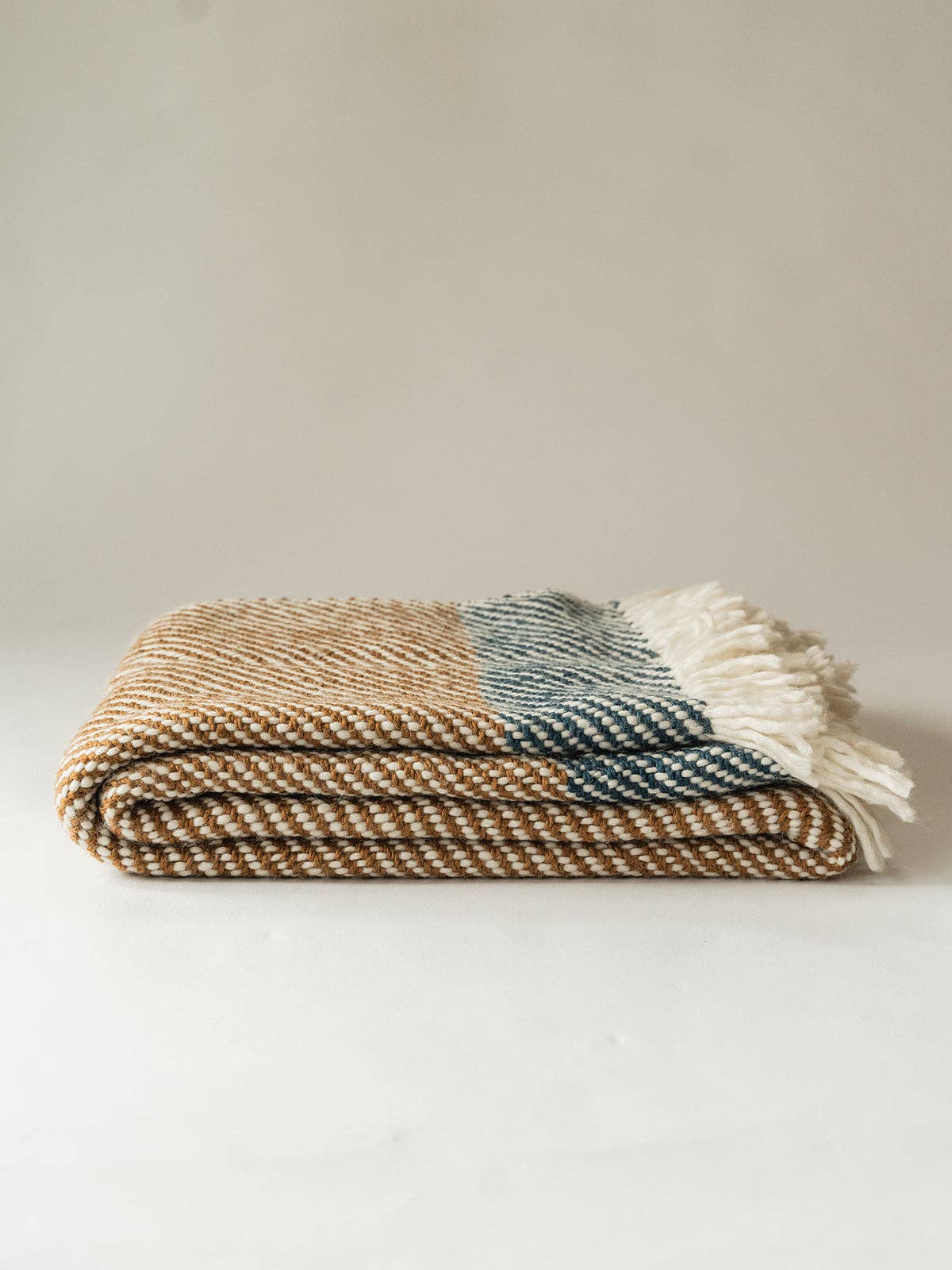 A folded woven throw blanket with a herringbone pattern in beige, brown, and muted blue-gray, finished with white fringe tassels, set against a plain light beige background.