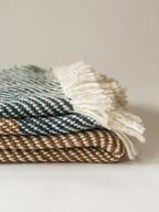 A folded woven throw blanket with cream fringe, shown in a close-up studio shot. The top visible section is blue and cream striped, and the folded layers below show a tan and cream pattern.