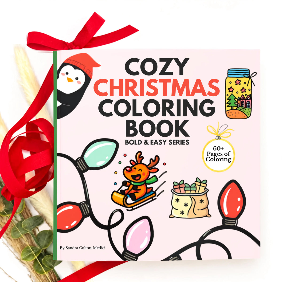 Cozy Christmas Coloring Book by Sandra Colton-Medici, featuring a light pink cover with festive illustrations of a penguin, a reindeer in a sled, Christmas lights, a gift sack, and a mason jar. The book is wrapped in a red ribbon and tied with a matching red bow, set against a styled background with dried grass and greenery.