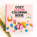 Cozy Easter Coloring Book from the Bold & Easy Series, featuring a light pink cover decorated with colorful Easter eggs, a bunny in a basket, and bold black and pink text, styled on a white surface with dried grass and green leaves.