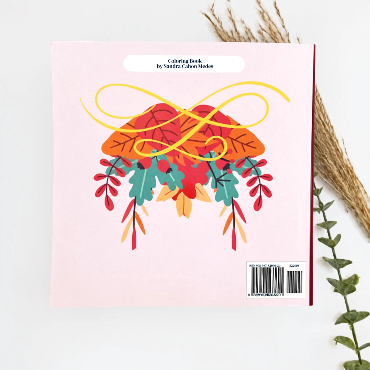 A coloring book by Sandra Colton-Medici shown from the back, featuring a light pink cover with a colorful autumn leaf illustration and a barcode label. The book is photographed on a white surface with dried grass and greenery as styled props.