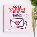 Cozy Valentine Coloring Book by Sandra Colton-Medici, featuring a light pink cover with bold text and an illustration of an envelope with a heart, styled on a white surface with dried wheat and green leaves.