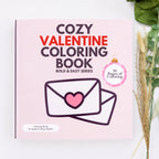 Cozy Valentine Coloring Book by Sandra Colton-Medici, featuring a light pink cover with bold text and an illustration of an envelope with a heart, styled on a white surface with dried wheat and green leaves.