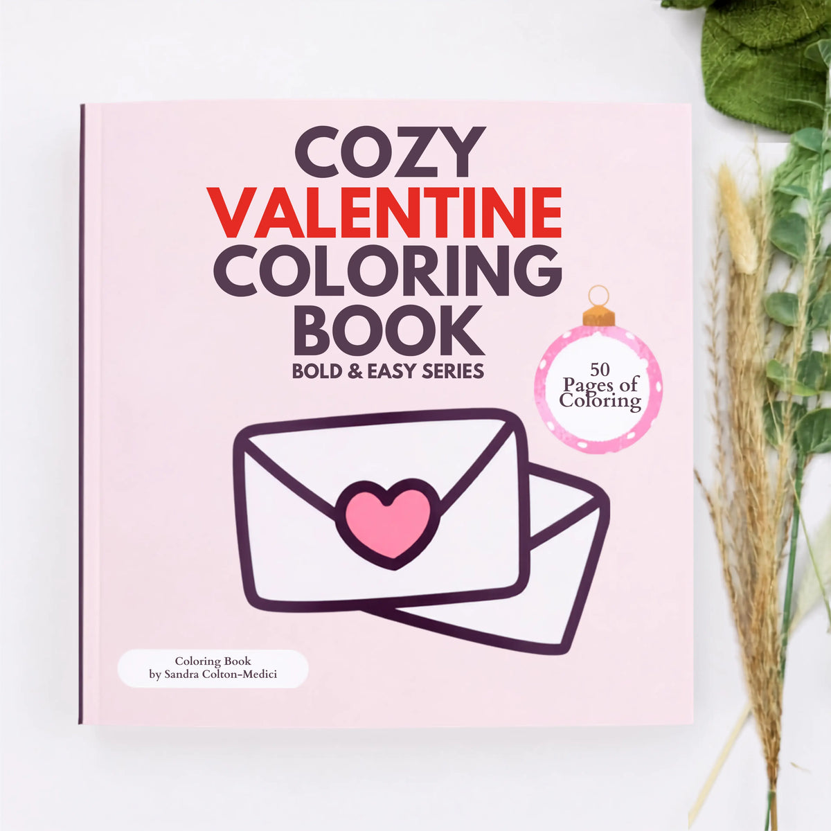 Cozy Valentine Coloring Book by Sandra Colton-Medici, featuring a light pink cover with bold text and an illustration of an envelope with a heart, styled on a white surface with dried wheat and green leaves.