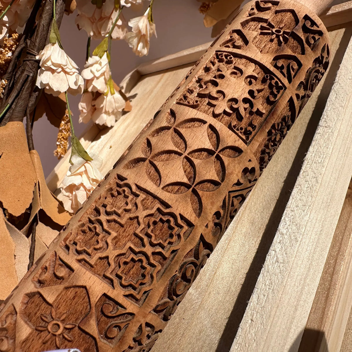 A close-up of a carved wooden rolling pin with intricate geometric and floral patterns, resting on a light-colored wooden surface with dried flowers in the background.