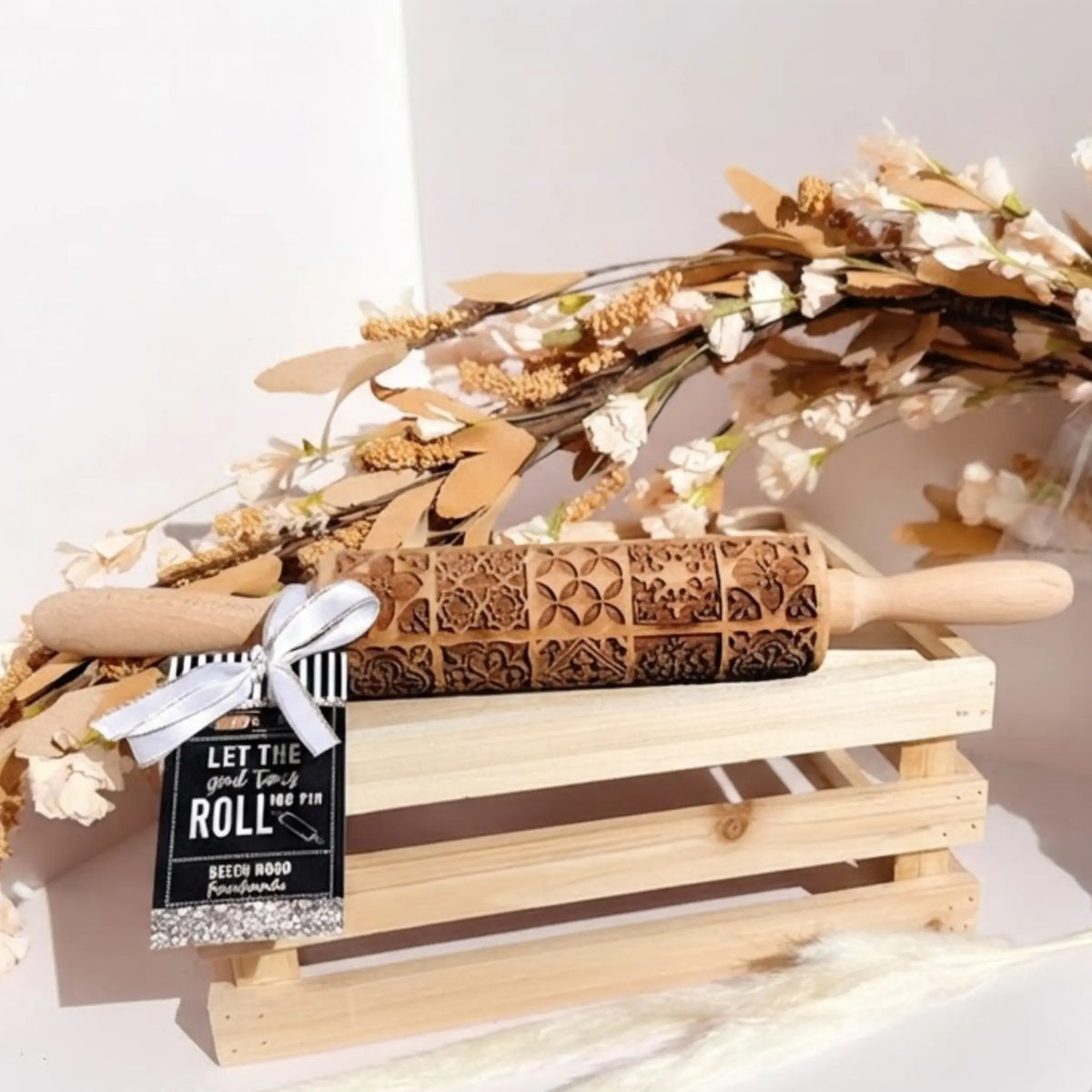 A carved wooden rolling pin with floral and geometric engravings, resting on a small wooden crate. A black-and-white striped ribbon and a hanging tag reading 'LET THIS good times ROLL be yours' are attached.