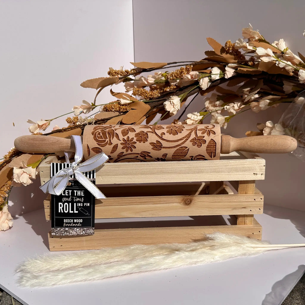 A carved beech wood rolling pin with floral engravings rests on a small wooden crate, tied with a white ribbon and a black tag that reads 'LET THE ROLL WIN.' Decorative dried flowers and a fluffy cream stem are arranged around it against a clean white background.