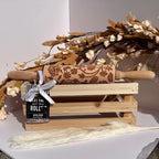 A carved beech wood rolling pin with floral engravings rests on a small wooden crate, tied with a white ribbon and a black tag that reads 'LET THE ROLL WIN.' Decorative dried flowers and a fluffy cream stem are arranged around it against a clean white background.