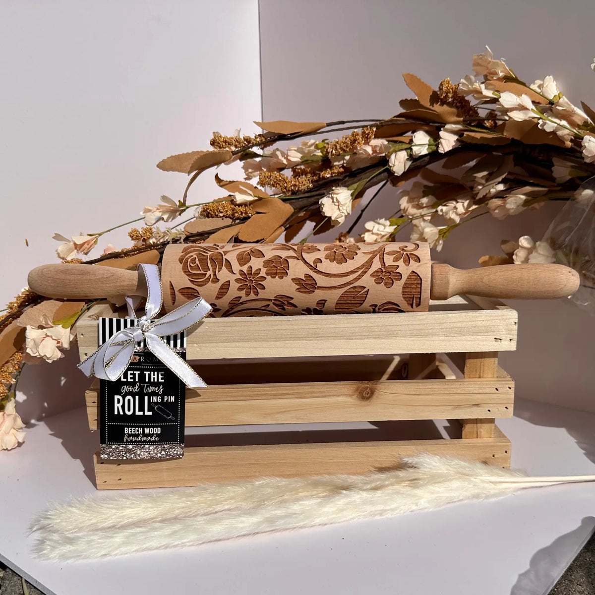 A carved beech wood rolling pin with floral engravings rests on a small wooden crate, tied with a white ribbon and a black tag that reads 'LET THE ROLL WIN.' Decorative dried flowers and a fluffy cream stem are arranged around it against a clean white background.