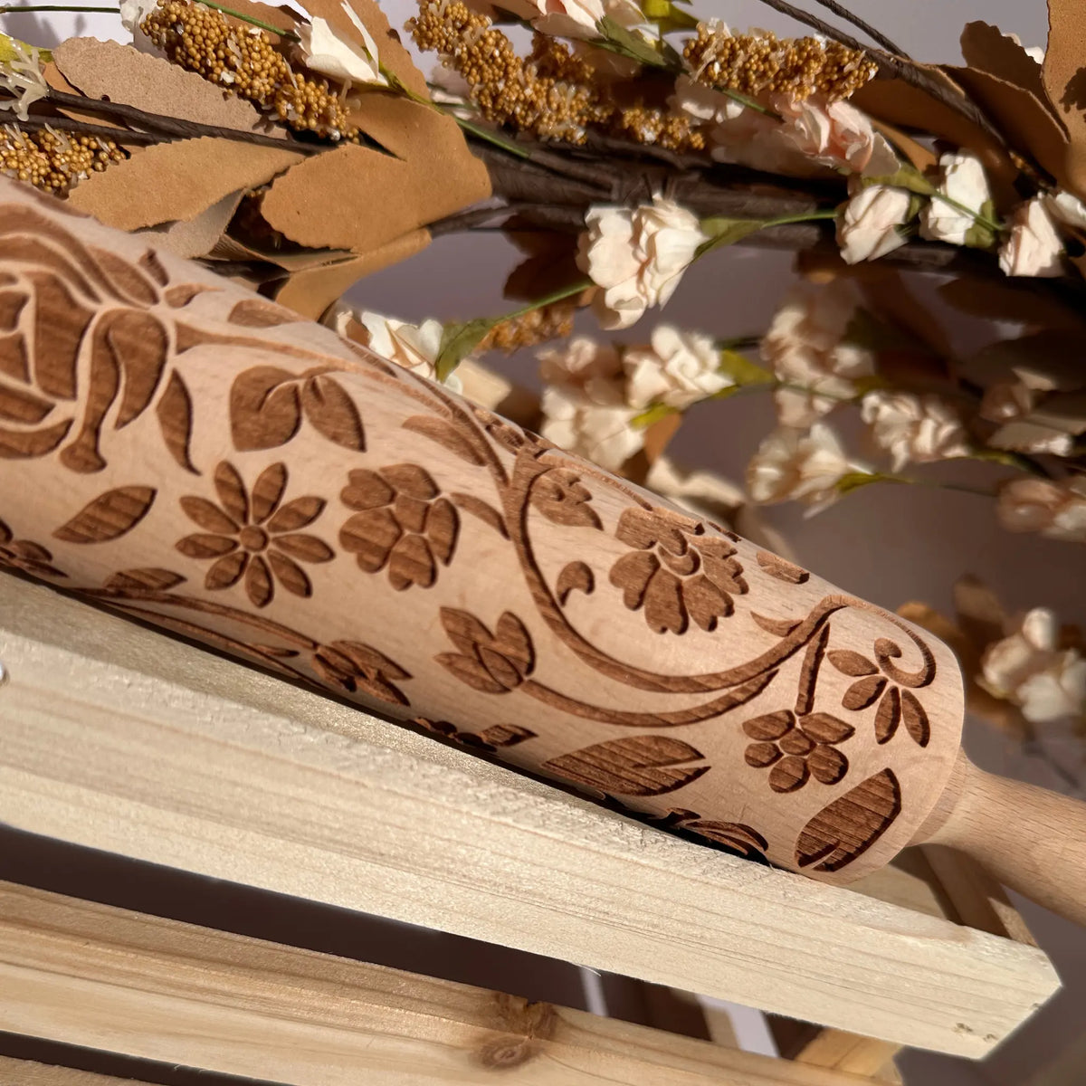 A close-up of a wooden rolling pin with an engraved floral pattern, resting on a light wooden surface with dried flowers in the background.