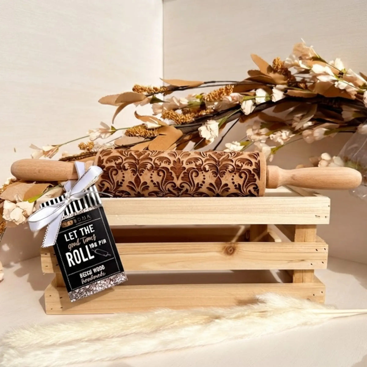 A wooden rolling pin with an engraved damask pattern and a black tag reading 'LET THE GOOD TIMES ROLL THE PIN' sits on a light wood crate with dried flowers and pampas grass.