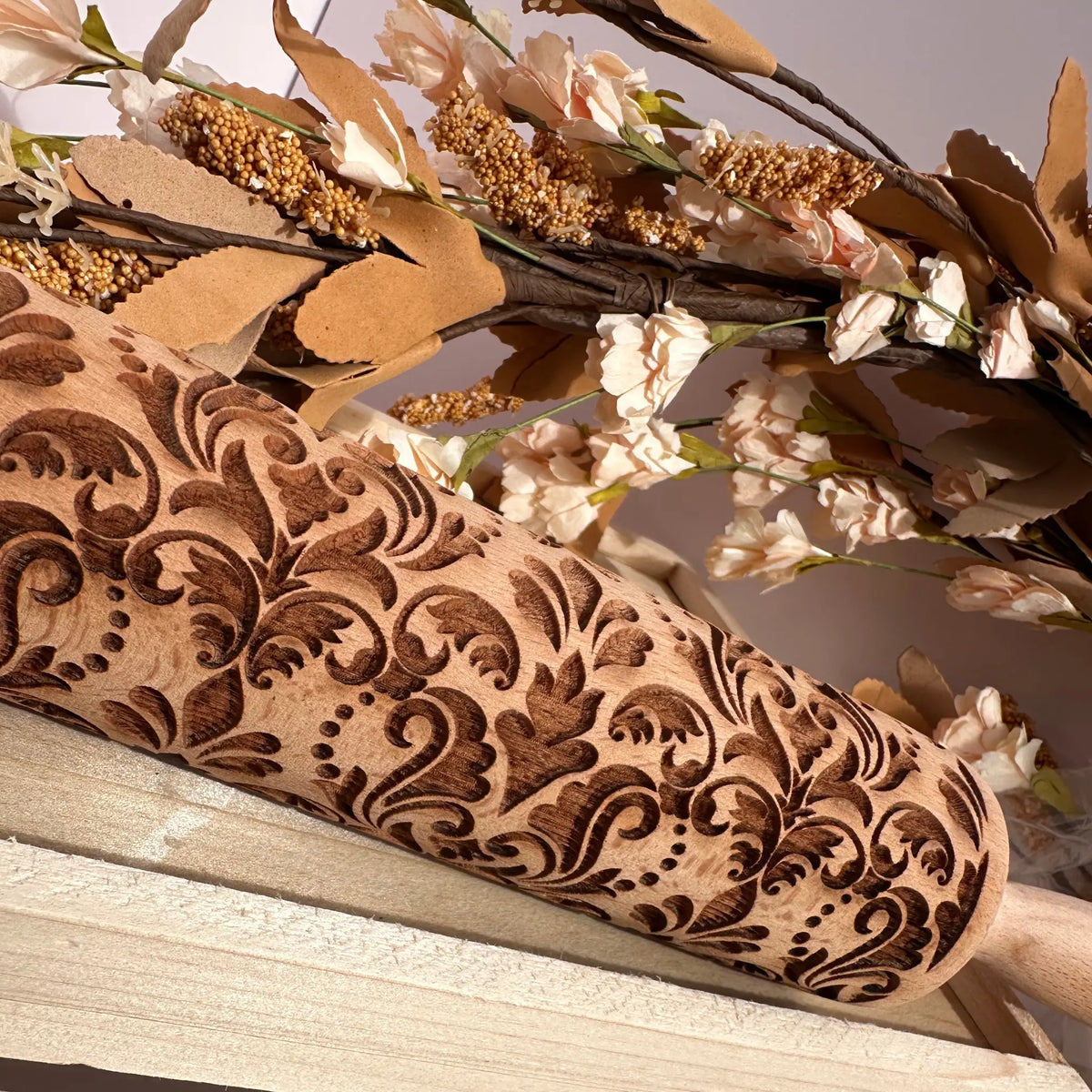 A close-up of a wooden rolling pin with an ornate, hand-carved damask pattern, resting on a light wooden surface with dried flowers and branches in the background.
