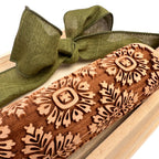 A close-up of a wooden rolling pin with laser-engraved floral and geometric patterns, resting on a light wood tray with an olive green ribbon bow.