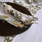 A styled tabletop setting featuring a white napkin with an olive green floral and bird toile pattern, wrapped in a beige rope napkin ring and placed on a dark brown wooden charger plate. A matching patterned tablecloth is visible in the background, decorated with dried bunny tail grass and greenery.