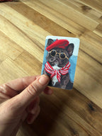 A hand holds a small illustrated card featuring a French bulldog wearing a red beret, round gold-rimmed sunglasses, and a red-and-white striped bow tie, against a light wood floor background.