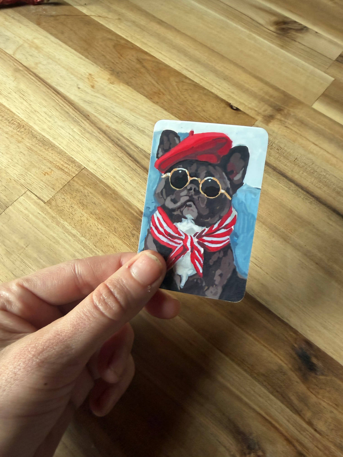 A hand holds a small illustrated card featuring a French bulldog wearing a red beret, round gold-rimmed sunglasses, and a red-and-white striped bow tie, against a light wood floor background.