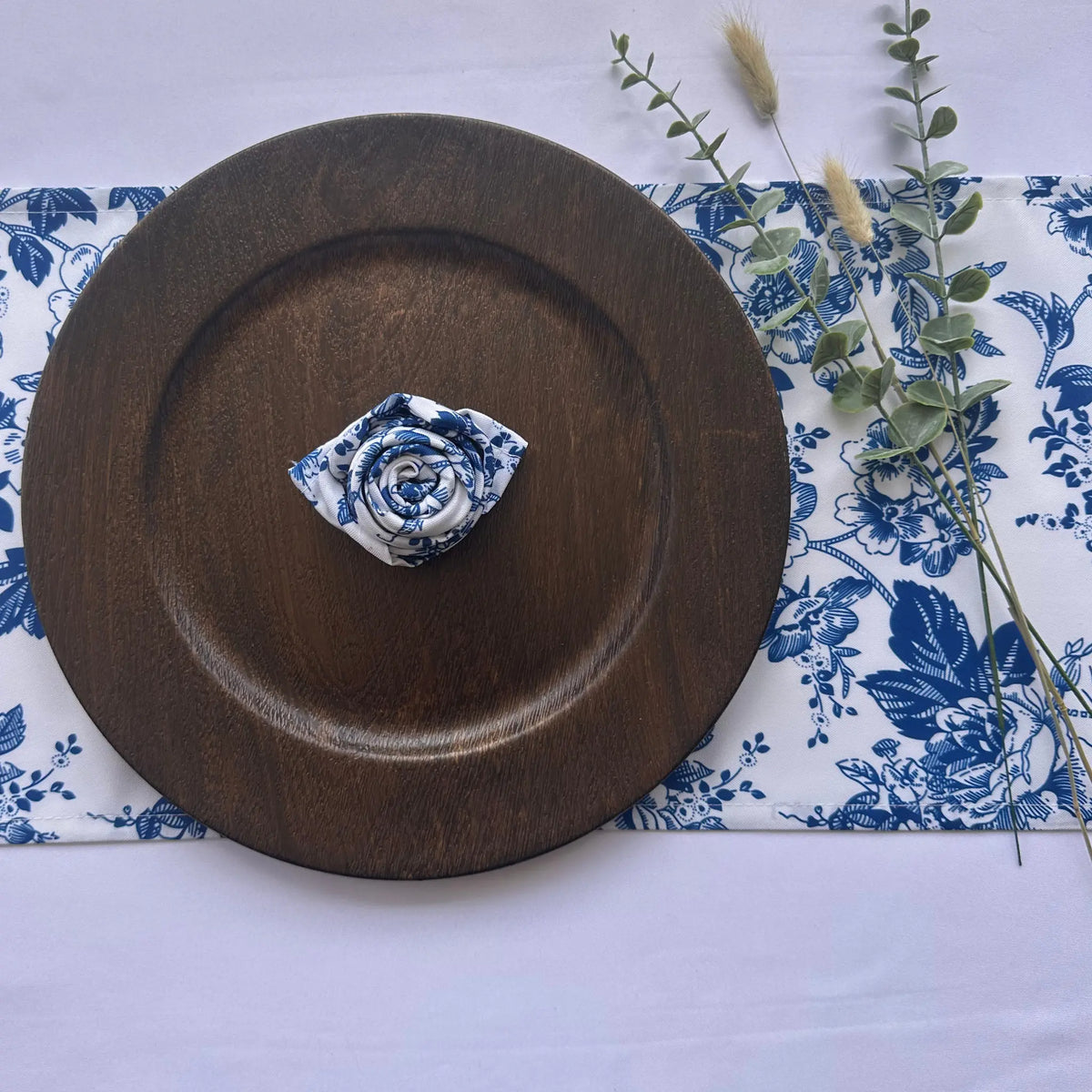 A blue and white floral fabric napkin folded into a rose shape, placed on a round dark brown wooden charger plate over a matching blue and white floral table runner with decorative eucalyptus stems.