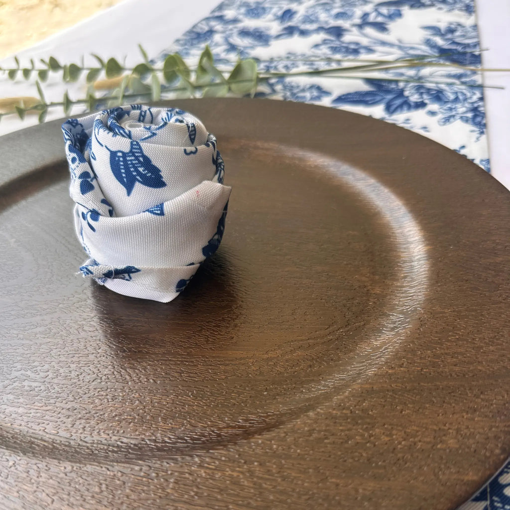A rolled white fabric napkin with a blue floral toile pattern, styled on a dark brown textured ceramic plate.