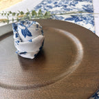 A rolled white fabric napkin with a blue floral toile pattern, styled on a dark brown textured ceramic plate.