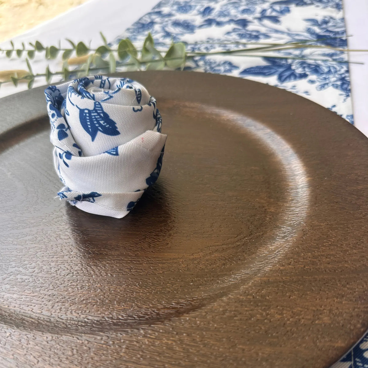 A rolled white fabric napkin with a blue floral toile pattern, styled on a dark brown textured ceramic plate.