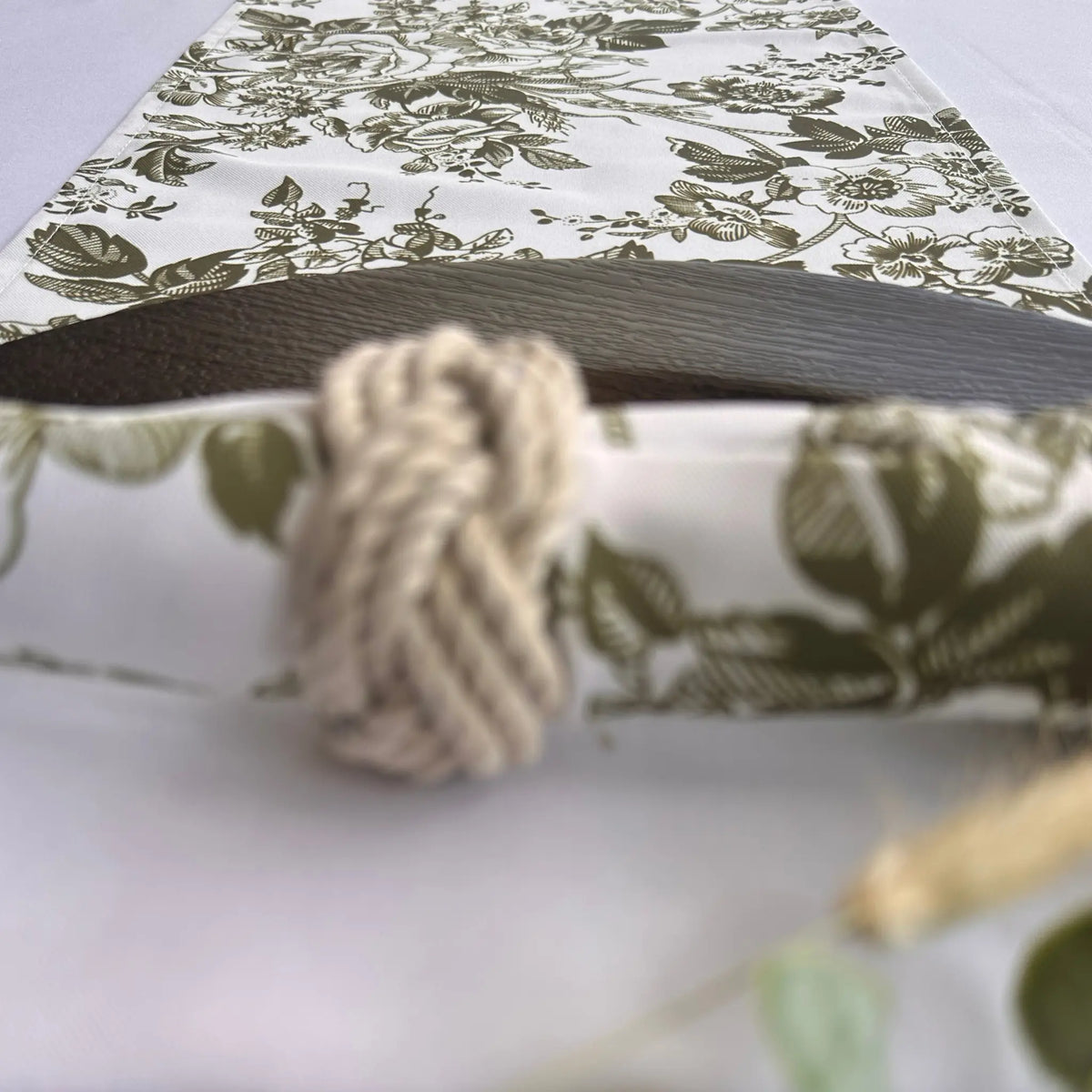 A close-up of a napkin with a white background and olive green floral toile pattern, featuring a rolled dark wood insert and a knotted beige rope detail.