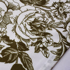 A close-up of a woven fabric with a dark olive-green floral toile pattern on a white background, showing the texture and a folded corner.
