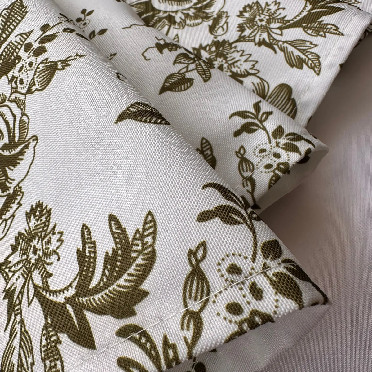 A close-up of folded fabric with a botanical toile-style print in olive green on a warm off-white background.