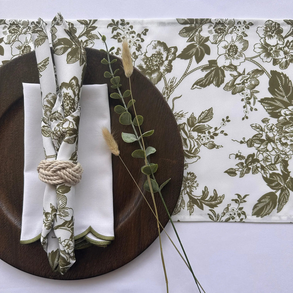 A styled tabletop setting featuring a white table runner and matching napkins with an olive green floral toile pattern, arranged on a dark brown wooden plate with a rope napkin ring and decorative grass stems.