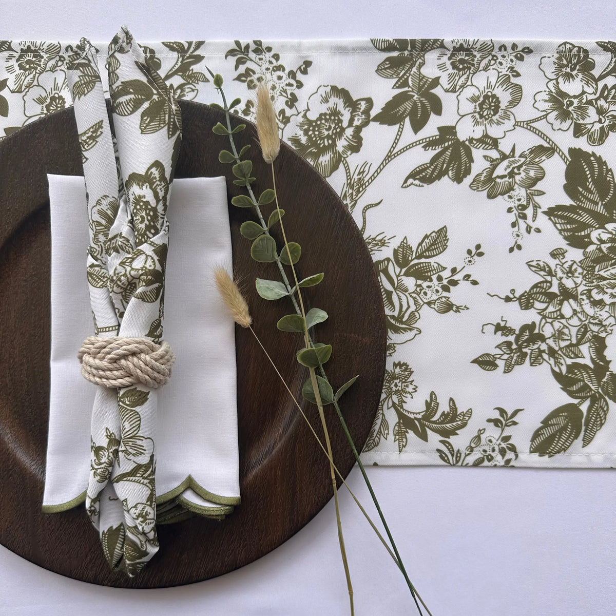 A styled tabletop setting featuring a white table runner and matching napkins with an olive green floral toile pattern, arranged on a dark brown wooden plate with a rope napkin ring and decorative grass stems.
