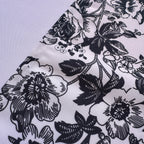 A close-up of a white fabric with a black floral and leaf pattern, laid diagonally across a light gray background.