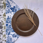 A round dark brown wooden plate with a shallow rim, styled on a white tablecloth with a blue floral napkin and a dried grass stem.