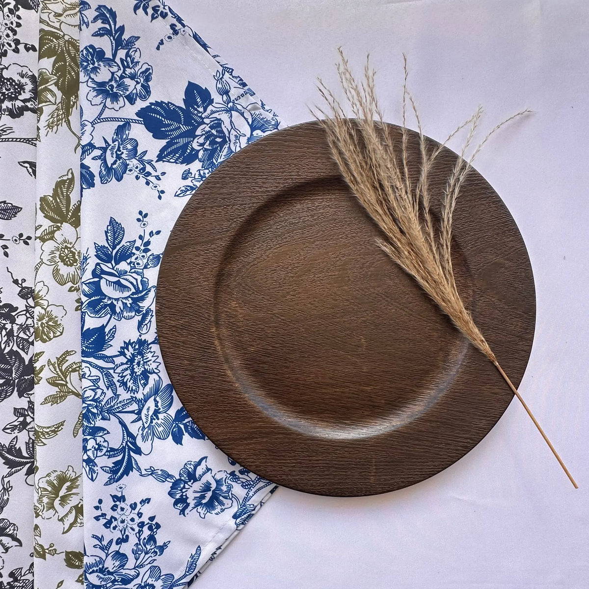 A round dark brown wooden plate with a shallow rim, styled on a white tablecloth with a blue floral napkin and a dried grass stem.