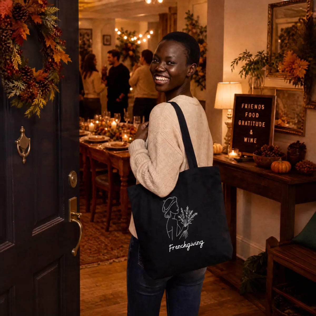 A woman wearing a cream knit sweater and jeans stands in a warmly lit dining room doorway, smiling while carrying a black canvas tote bag. The bag has a white line drawing of a woman and the word 'Frenchgiving' printed on the front.