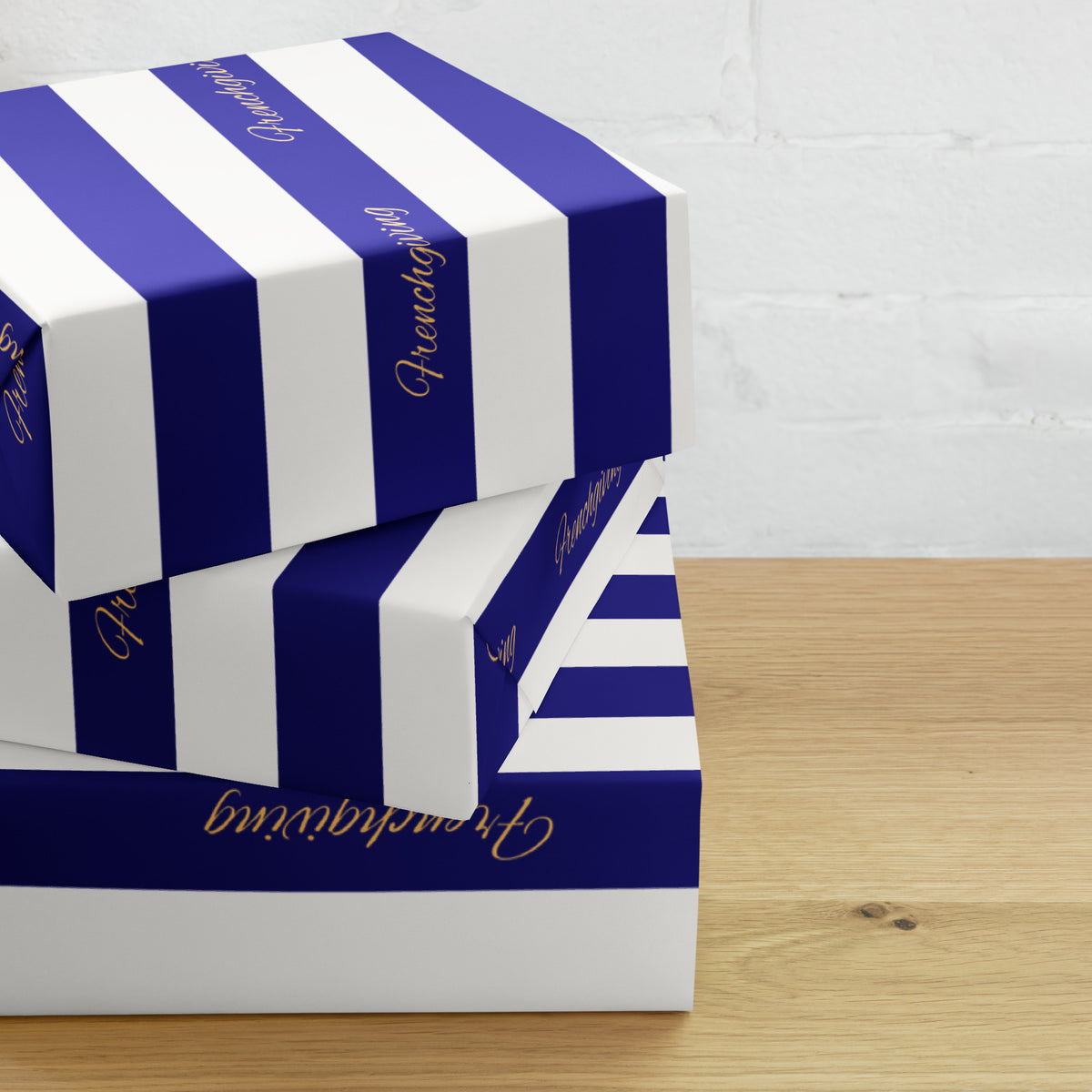 Three stacked gift boxes with bold royal blue and white vertical stripes, accented with gold foil script text, set on a light wood tabletop against a white brick wall.