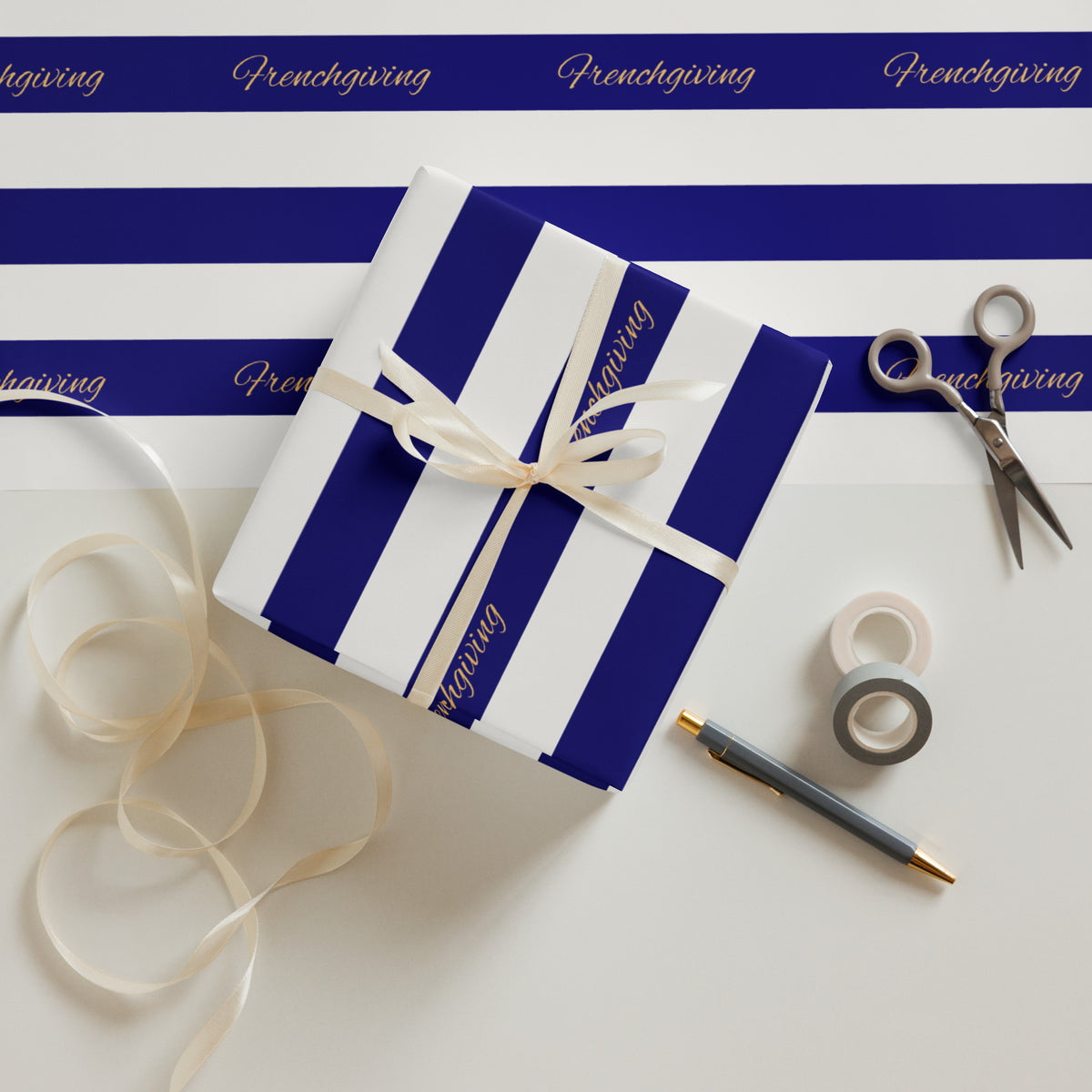 A wrapped gift box with bold royal blue and white vertical stripes, tied with a cream ribbon, arranged on a white surface with matching striped paper, scissors, washi tape, a pen, and loose ribbon. The paper shows the word 'Frenchgiving' repeated across it.