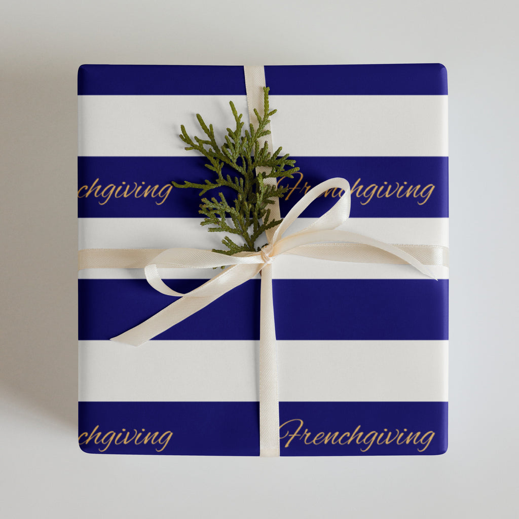 A square gift box wrapped in royal blue and white striped paper with the word 'Frenchgiving' printed in gold script. The box is tied with a cream ribbon and topped with a small green pine sprig, photographed against a light gray background.