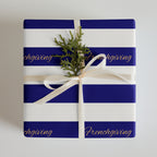 A square gift box wrapped in royal blue and white striped paper with the word 'Frenchgiving' printed in gold script. The box is tied with a cream ribbon and topped with a small green pine sprig, photographed against a light gray background.
