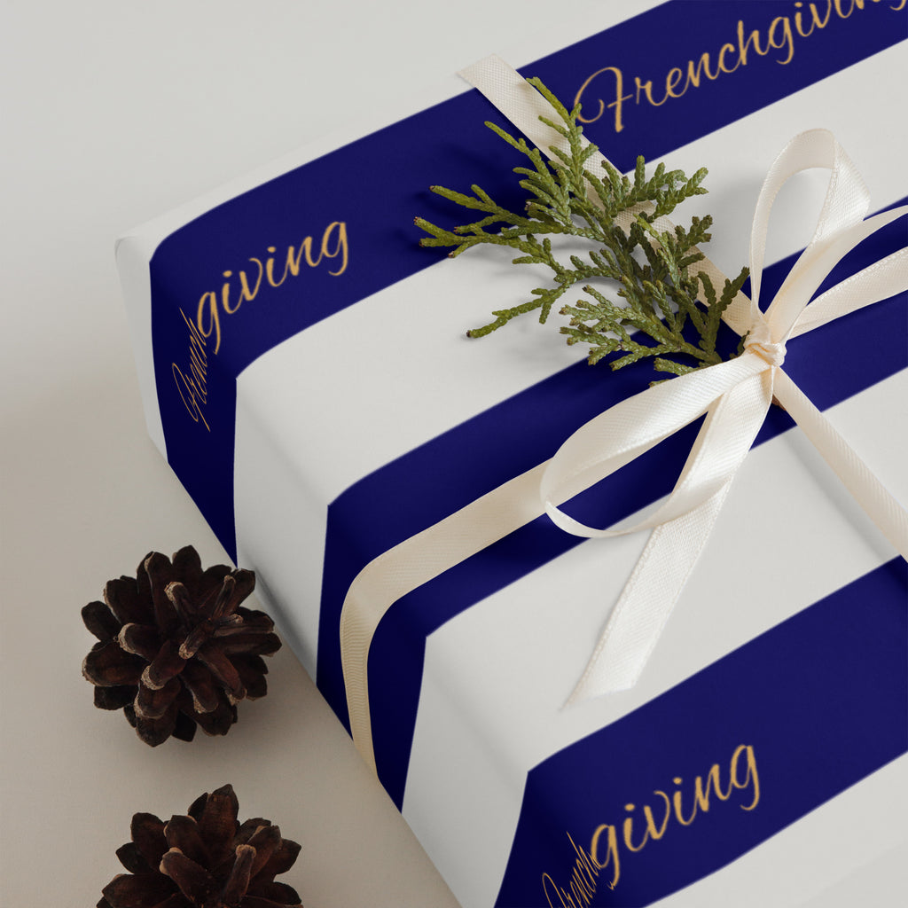 A gift box wrapped in white paper with bold royal blue stripes and gold script reading 'Frenchgiving,' tied with a cream ribbon and topped with a small green pine sprig, with pinecones on a light neutral background.