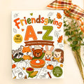Friendsgiving A-Z Kids Color & Trace Activity Book cover displayed on a light gray surface with a plaid ribbon and dried grasses as styling props.
