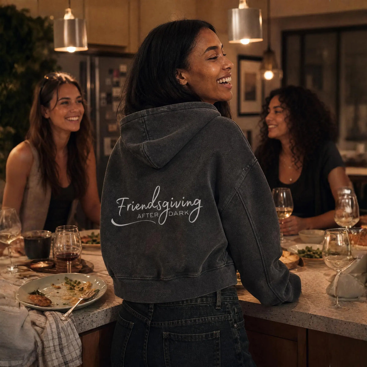 A woman standing at a kitchen island in a warmly lit dining/kitchen setting, wearing a dark charcoal gray hoodie with 'Friendsgiving AFTER DARK' printed in white script across the back. Two other women are seated at the table in the background.