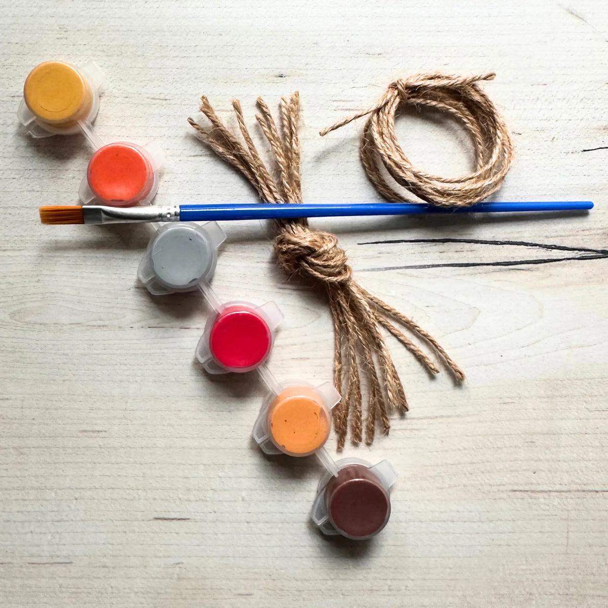 A DIY craft kit featuring natural jute twine, a blue-handled paintbrush, and six small plastic paint pots in yellow, orange, red, gray, orange, and brown, arranged on a light wood background.